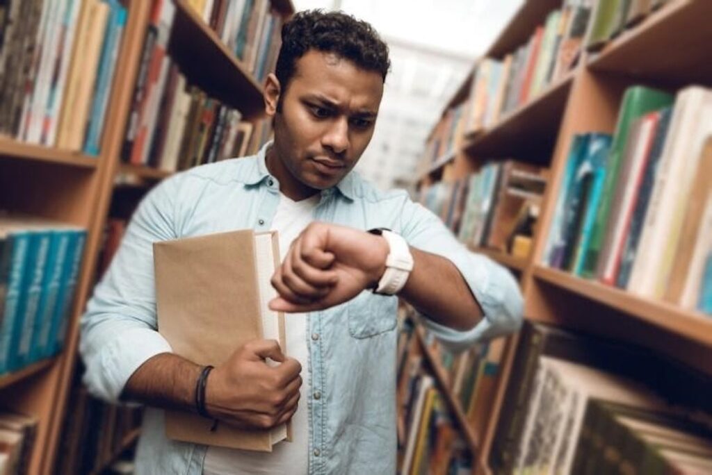 A student in an aisle of a library building’s stacks holds a textbook while staring at their watch.