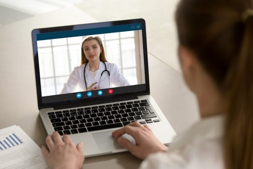 A person with a laptop has a virtual conference with a person wearing a white physician’s coat.