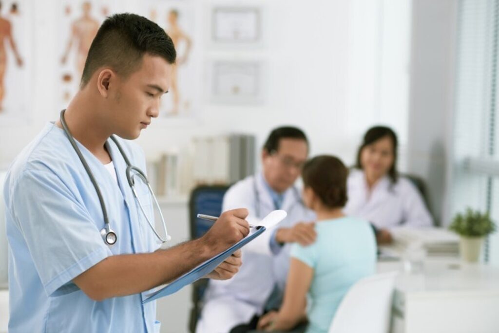 A person in scrubs with a stethoscope writes on a pad while people in a clinic receive health care.