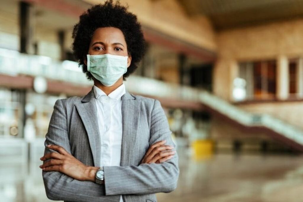A manager in business attire wearing a face mask stands in a hospital corridor with arms folded.