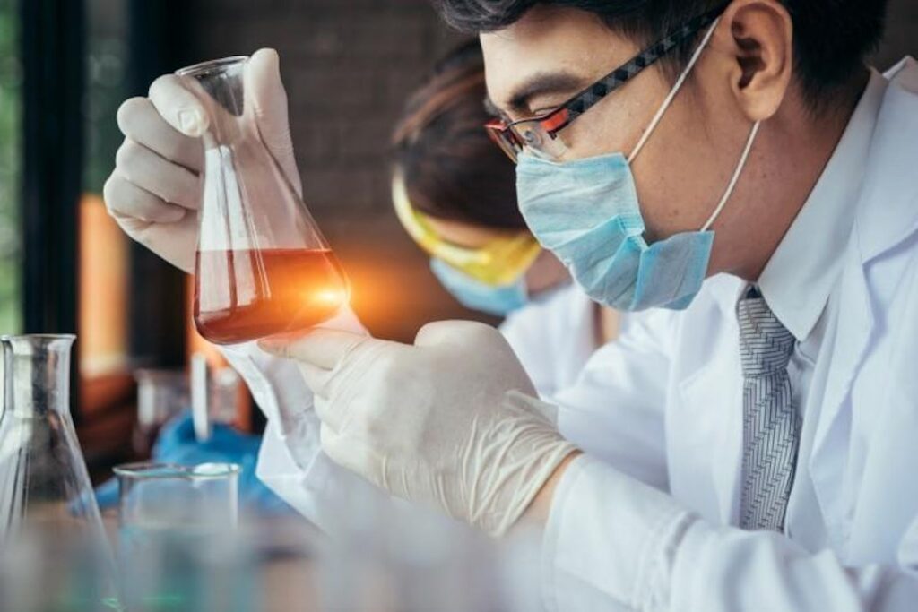 In a lab, a person in a white coat and protective gear examines the contents of a beaker.