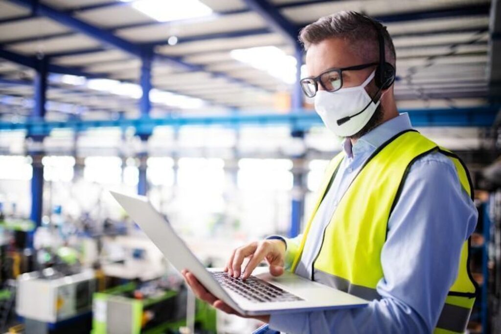 A person wearing a protective mask and a headset holds a laptop in an industrial factory.