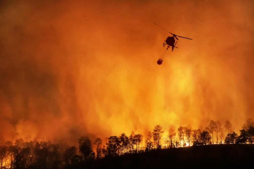 A helicopter carries a water bucket over a wildfire.