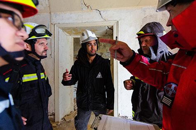 A group of first responders wearing emergency gear and hard hats stand in a circle inside a room with cracked walls