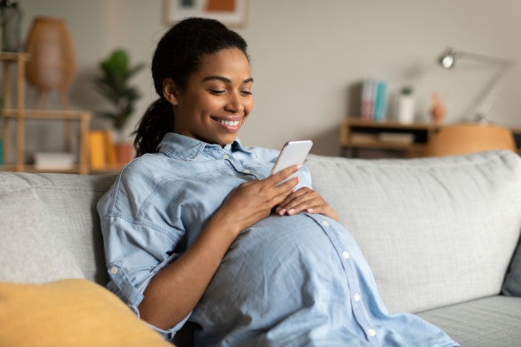 A smiling pregnant individual sits at home looking at a phone.