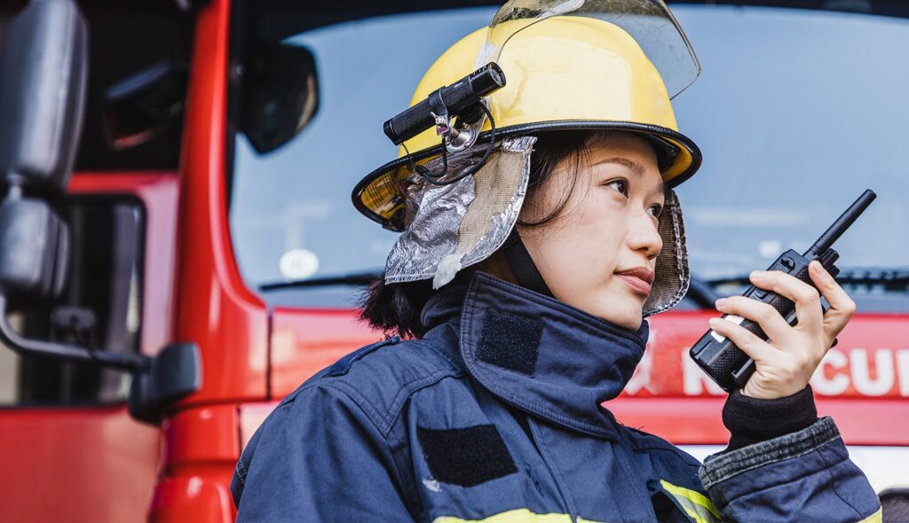 A disaster management professional wearing a helmet and bunker coat listens to a walkie-talkie they hold in her hand.